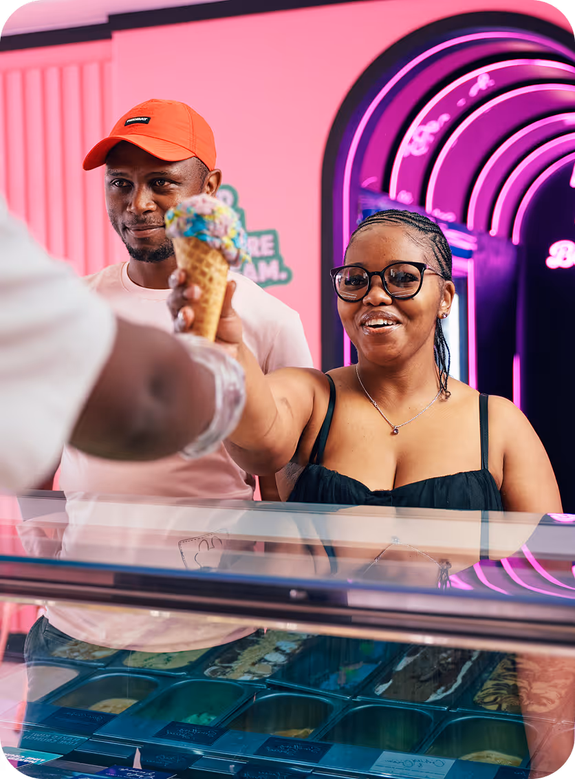Woman with glasses and braided hair smiling as she receives a colorful ice cream cone from a vendor, with a man in an orange cap standing beside her.
